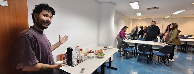 A man in a purple shirt stands in a room with a table and chairs. Description generated by AI