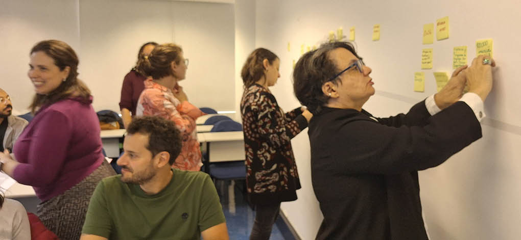 A group of people are standing in front of a white wall, writing on it with yellow sticky notes. Description generated by AI